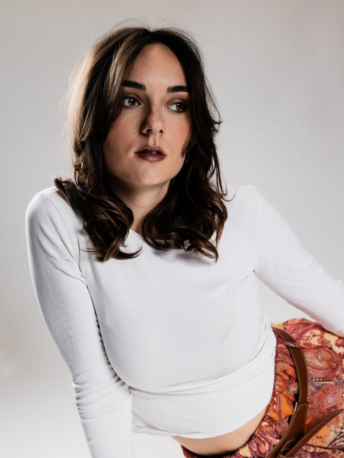 Portrait en studio d'une jeune femme brune aux cheveux mi-longs ondulés, en haut blanc à manches longues, sur fond clair.
